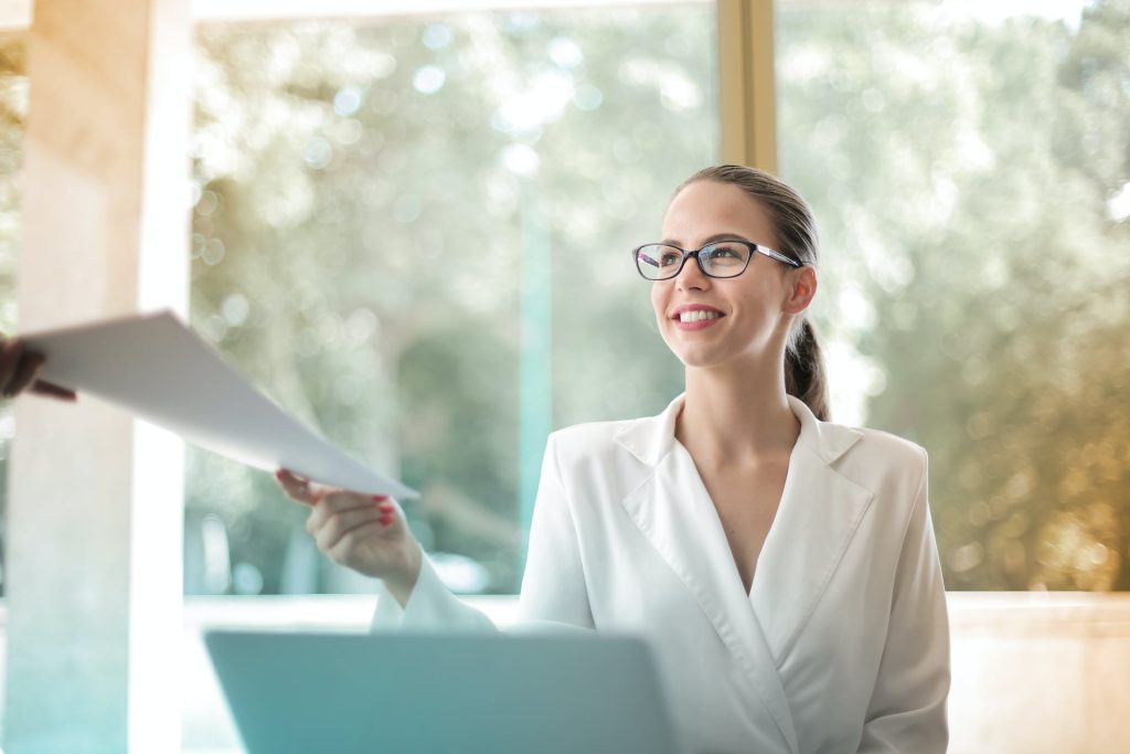 Low angle of successful female executive manager in classy style sitting at table with laptop in contemporary workplace and passing documents to colleague